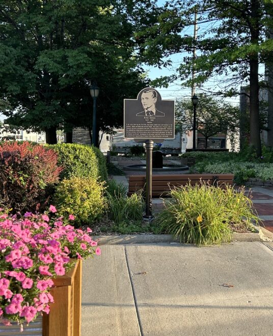 Memorial Plaque: Bronze historical marker for Jesse James displayed in a landscaped park with benches, greenery, and flowering plants along a brick path and concrete sidewalk.