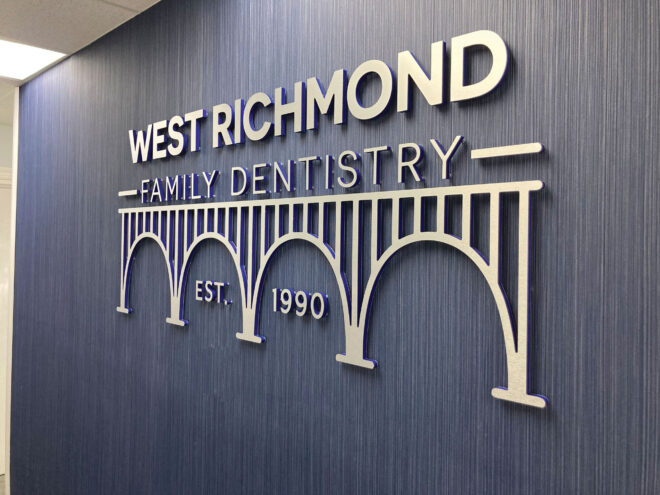 Laminate – Interior (West Richmond Family Dentistry): Laminate dimensional letters mounted behind a reception desk for West Richmond Family Dentistry. Interior lobby signage is installed on a blue painted wall with soft lighting.
