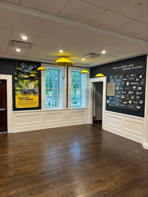 Bright modern interior with branded wall graphics, alumni display wall, yellow pendant lights, and hardwood flooring