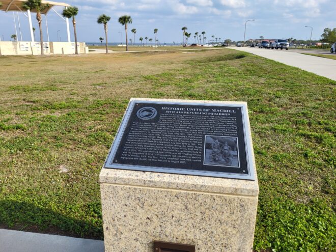 Airforce Plaque: Plaque titled ‘Historic Units of MacDill – 50th Air Refueling Squadron’ mounted on a stone pedestal beside a wide grassy field with palm trees and a road lined with cars under a partly cloudy sky.